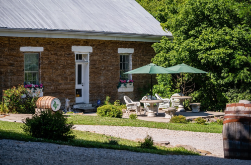 A rustic stone building with a gray roof, surrounded by greenery, featuring outdoor seating and umbrellas.