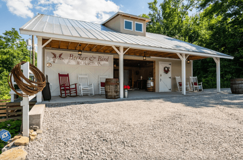 A rustic building with a sign reading "Berker & Bird Bourbon," featuring rocking chairs and barrels outside.
