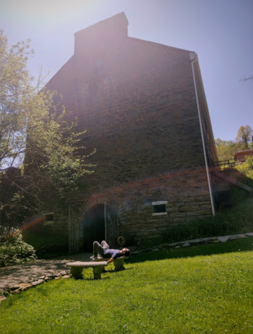 A person lies on a stone bench in front of a large, old stone building surrounded by greenery under a bright sky.