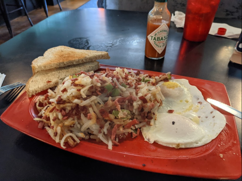 A red plate with hash browns mixed with meat, two fried eggs, and toast, alongside a bottle of Tabasco sauce.