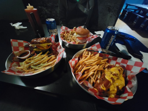 Three plates of food featuring burgers, fries, and onion rings, with two cans of Bud Light on a table.