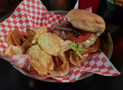 A burger with lettuce, tomato, and cheese served with a side of seasoned potato chips in a basket with red checkered paper.