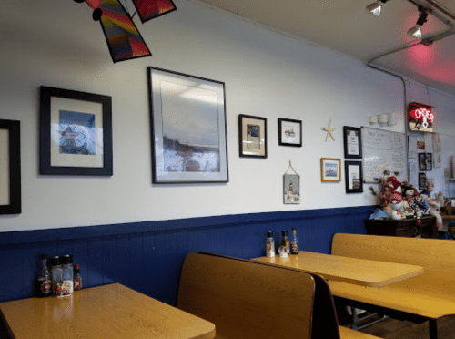 A cozy diner interior with wooden tables, framed artwork on the walls, and decorative items, including a neon sign.
