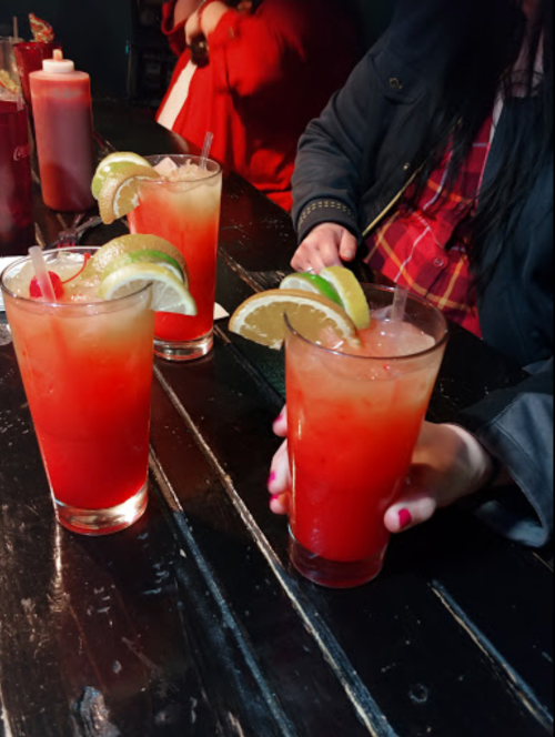 Three colorful cocktails with lime and lemon slices on a dark wooden table, with a hand holding one drink.