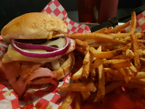A burger with cheese, ham, onions, and pickles, served with a side of crispy French fries on a red checkered tray.