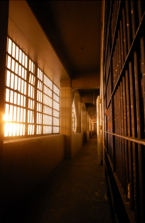 A dimly lit prison corridor with barred windows casting warm light on the walls.