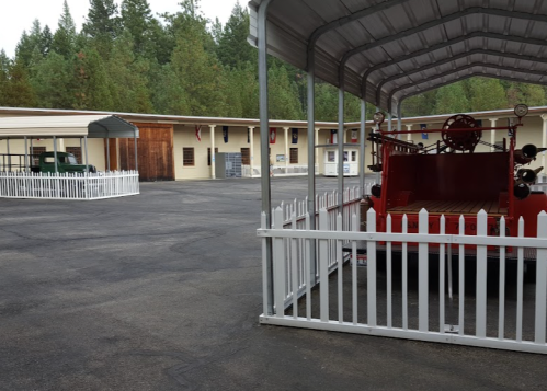 A parking area with a vintage fire truck under a shelter, surrounded by a white picket fence and trees in the background.