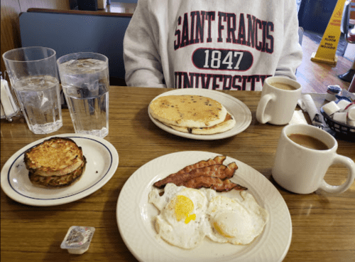 A breakfast spread with pancakes, eggs, bacon, a sandwich, coffee, and water at a diner table.