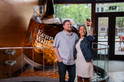 A man and woman take a selfie in front of a large bourbon glass display in a bar setting.