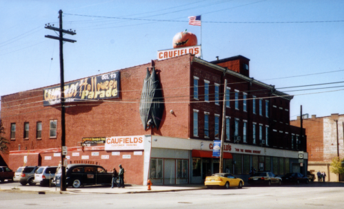A brick building with a large pumpkin sign, featuring storefronts and parked cars on a sunny day.