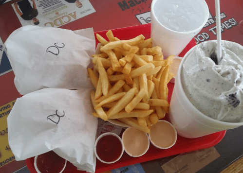 A red tray with two wrapped sandwiches labeled "DC," a pile of fries, and two cups of drinks alongside dipping sauces.
