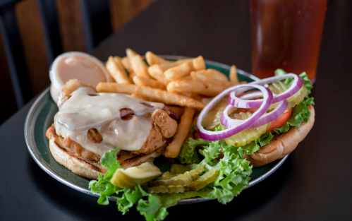 A plate with a chicken sandwich topped with cheese, fries, and a side of lettuce, tomato, onion, and pickles.