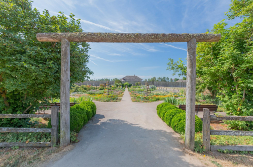 A wooden archway frames a pathway leading to a garden with lush greenery and a distant building under a blue sky.