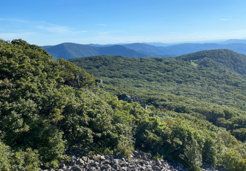 A panoramic view of lush green mountains under a clear blue sky, with rocky outcrops in the foreground.