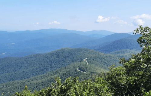A panoramic view of rolling green mountains under a clear blue sky, with a winding road visible in the distance.