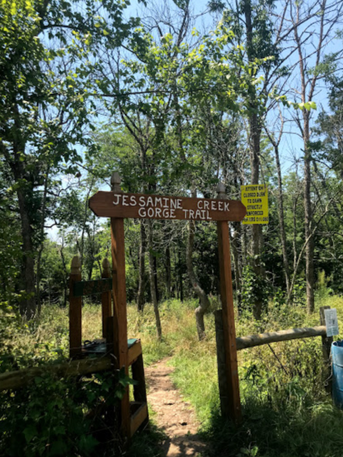 Wooden sign for Jessamine Creek Gorge Trail surrounded by trees and greenery on a sunny day.
