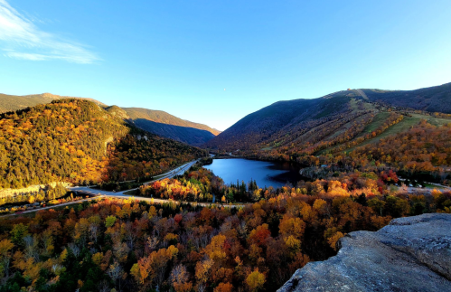 A scenic view of a lake surrounded by mountains and vibrant autumn foliage under a clear blue sky.