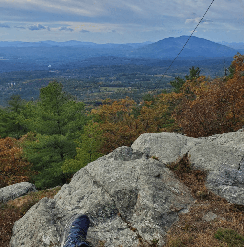 A view from a rocky ledge overlooking a valley with mountains in the distance, surrounded by autumn foliage.