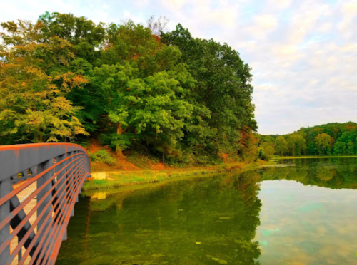 A serene view of a bridge over a calm lake, surrounded by lush trees and a colorful sky.