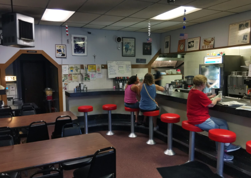 A cozy diner interior with red stools, two patrons at the counter, and tables in the background. Decor includes posters and flags.