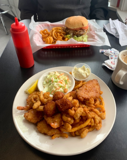 A plate of fried seafood with coleslaw, tartar sauce, and a lemon wedge, alongside a burger and curly fries in the background.