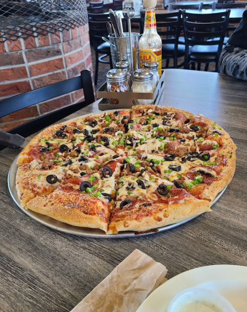 A large pizza topped with pepperoni, sausage, olives, and green onions on a wooden table in a restaurant setting.