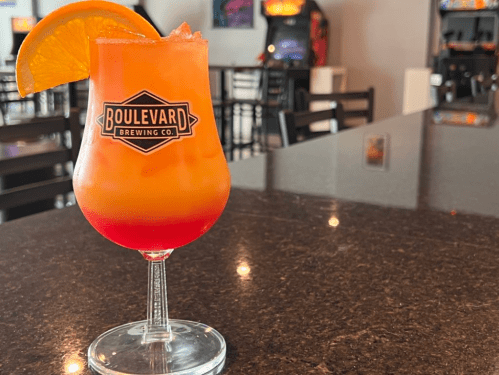 A colorful cocktail in a glass with an orange slice, sitting on a dark table in a bar setting.