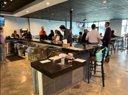 A busy bar area with patrons socializing, seated at high tables, and a bartender serving drinks.