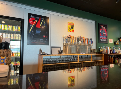 A bar with a mirrored countertop, colorful posters on the wall, and a row of beer taps behind the counter.