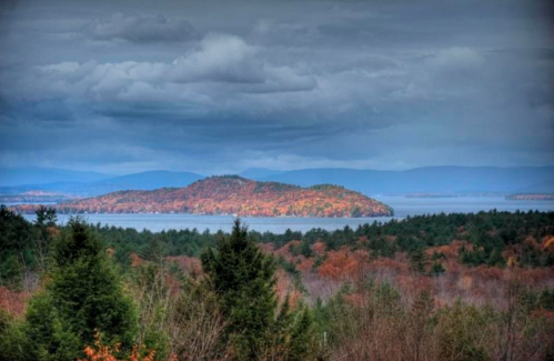 A scenic view of a lake surrounded by autumn-colored trees and distant mountains under a cloudy sky.