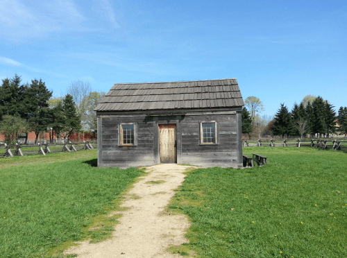 A small, weathered wooden house on a grassy field with a dirt path leading to the door, surrounded by trees.