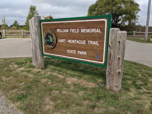 Sign for William Field Memorial Hart-Montague Trail at State Park, featuring wooden posts and grassy surroundings.