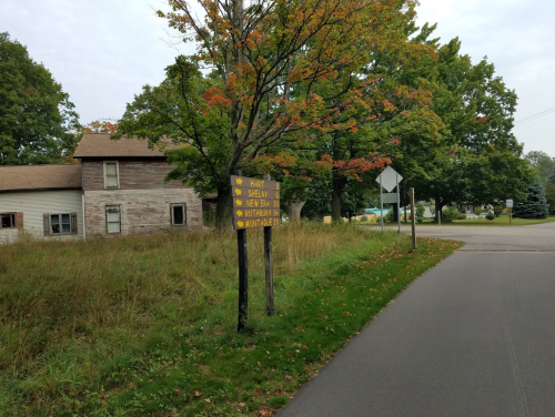 A weathered signpost with directional arrows for nearby towns, surrounded by overgrown grass and trees in autumn colors.