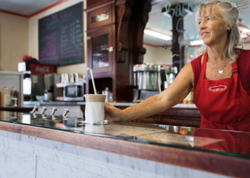 A woman in a red apron serves a milkshake at a vintage-style diner with a marble counter.