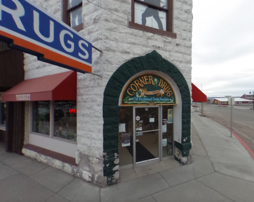 Exterior of a vintage drugstore with a green archway, featuring a sign that reads "Corner Drug" and nearby shops.