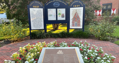 A historical marker and informational boards surrounded by flowers in a park setting, with a brick pathway.