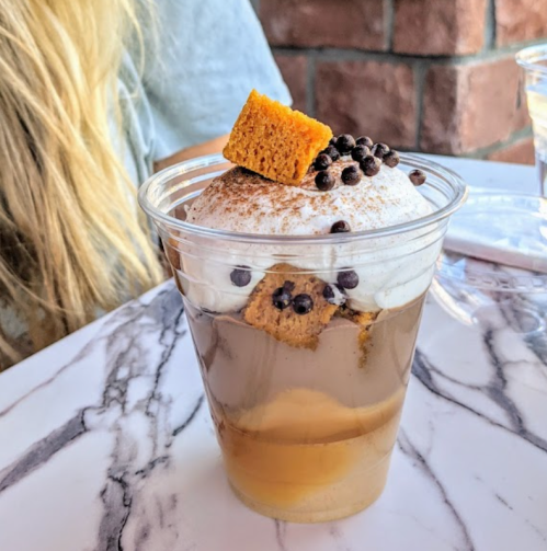 A clear cup filled with layered dessert, featuring coffee, cream, and topped with honeycomb and chocolate sprinkles.