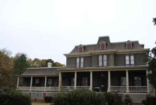 A large, two-story Victorian-style house with a wraparound porch and ornate details, surrounded by trees.