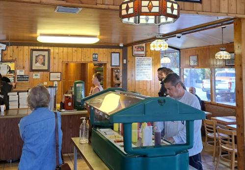 A cozy diner interior with wooden walls, customers at a salad bar, and a staff member serving food.