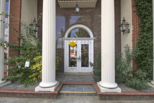 A grand entrance with white double doors, flanked by tall columns and greenery, featuring a decorative wreath.