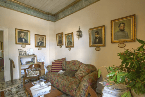 A cozy room with a floral couch, a wooden table, and framed portraits on the walls, featuring a potted plant.