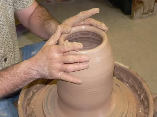 A person shaping a clay pot on a pottery wheel, hands covered in wet clay, focused on the creation process.