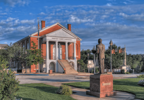 A historic courthouse with a statue in front, surrounded by greenery and a clear blue sky.