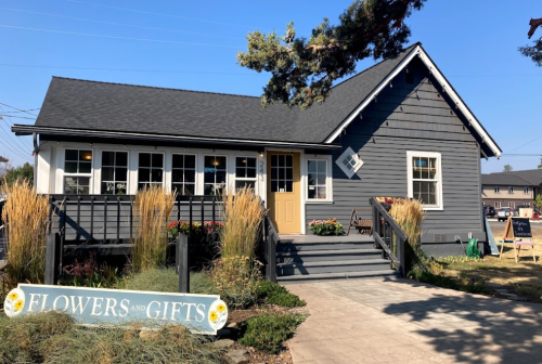 A charming gray house with a yellow door, featuring a "Flowers and Gifts" sign and colorful plants in front.
