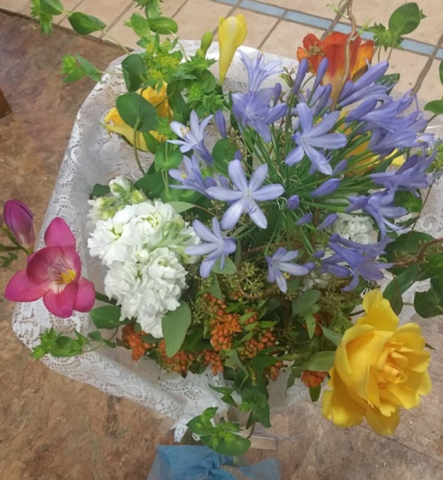 A vibrant bouquet featuring purple, yellow, pink, and white flowers, arranged with greenery and lace accents.