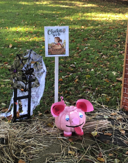 A pink pumpkin shaped like a pig sits on hay, next to a sign for "Charlotte's Web" and a spider web decoration.