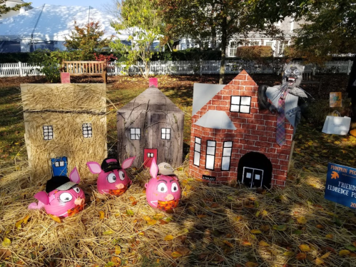 A whimsical Halloween display with straw, painted pig pumpkins, and cardboard houses, featuring a spooky figure.