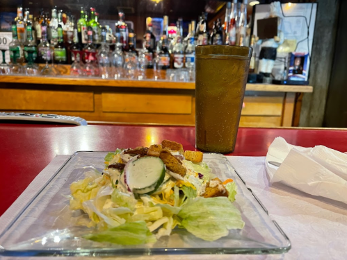 A glass of drink next to a salad on a glass plate, with a bar and bottles in the background.