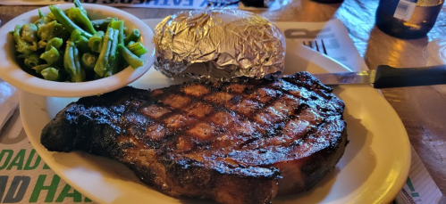 A grilled ribeye steak with a baked potato wrapped in foil and a side of green beans on a plate.