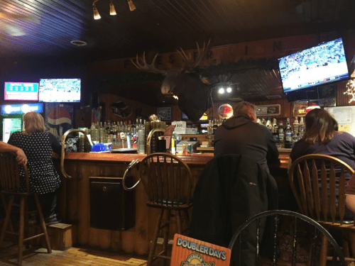A cozy bar with wooden decor, a moose head on the wall, and patrons sitting at the counter watching sports on TV.